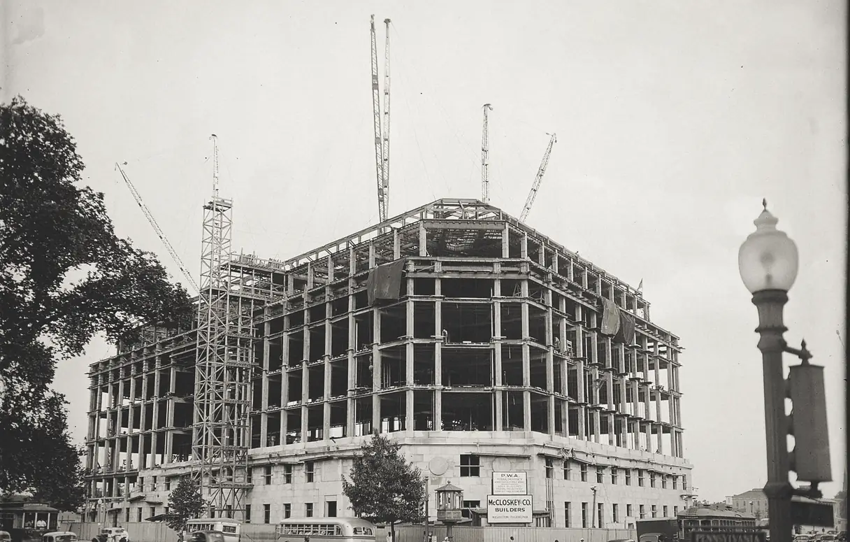 Steel frame of a large Washington office building under construction at a street corner, with four cranes above it, a stone base with a builder's sign, cars, buses and a streetcar in front, and a street lamp on the right.