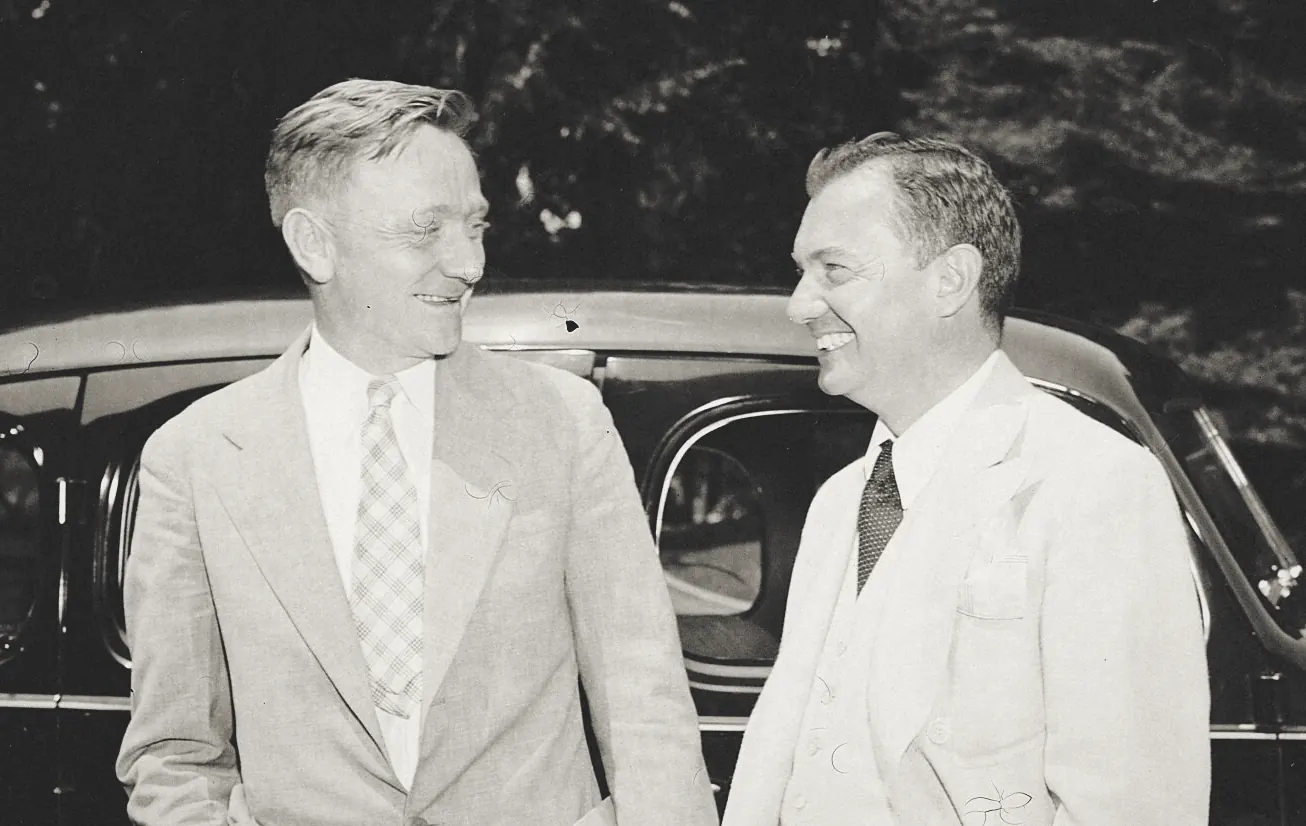 Two smiling men in light summer suits standing close and talking in front of a dark car, with trees behind them.